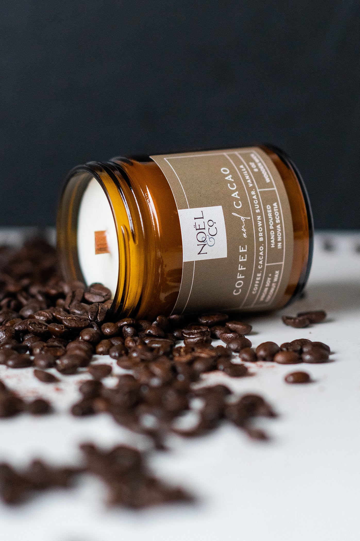 A candle in an amber glass jar and coffee beans on the table around it.