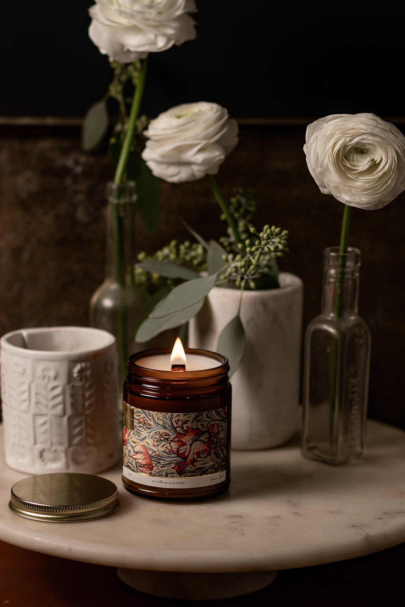 A lit candle in an amber glass jar with a wooden wick and a original art piece featured on the label.