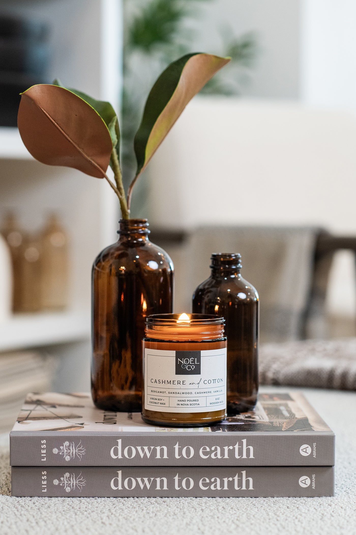 A lit candle in an amber glass jar with a wooden wick on two books next to two decorative amber glass bottles, one with a plant inside.