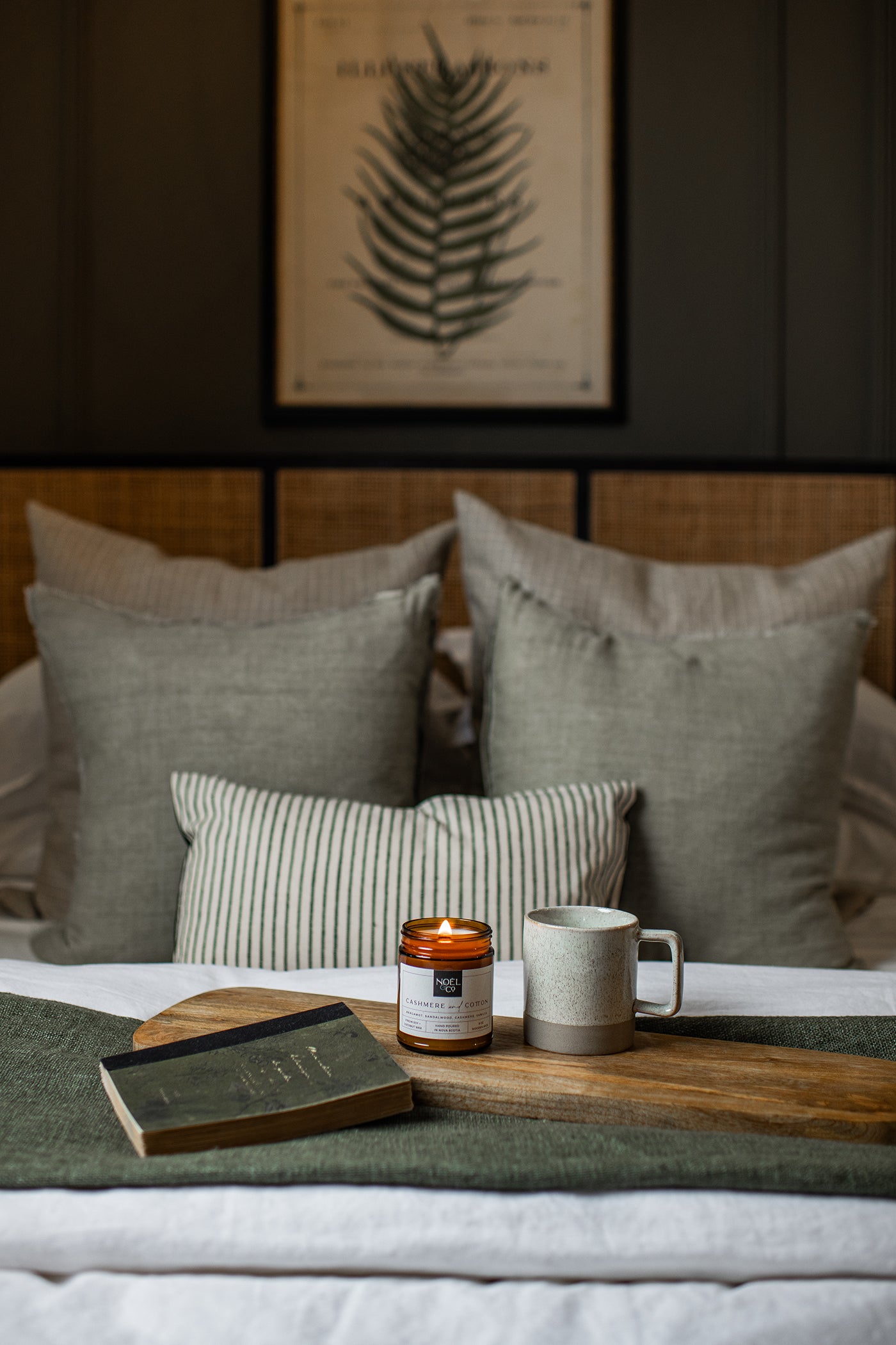 A lit candle in an amber glass jar with a wooden wick on a wooden slab next to a book and a mug.