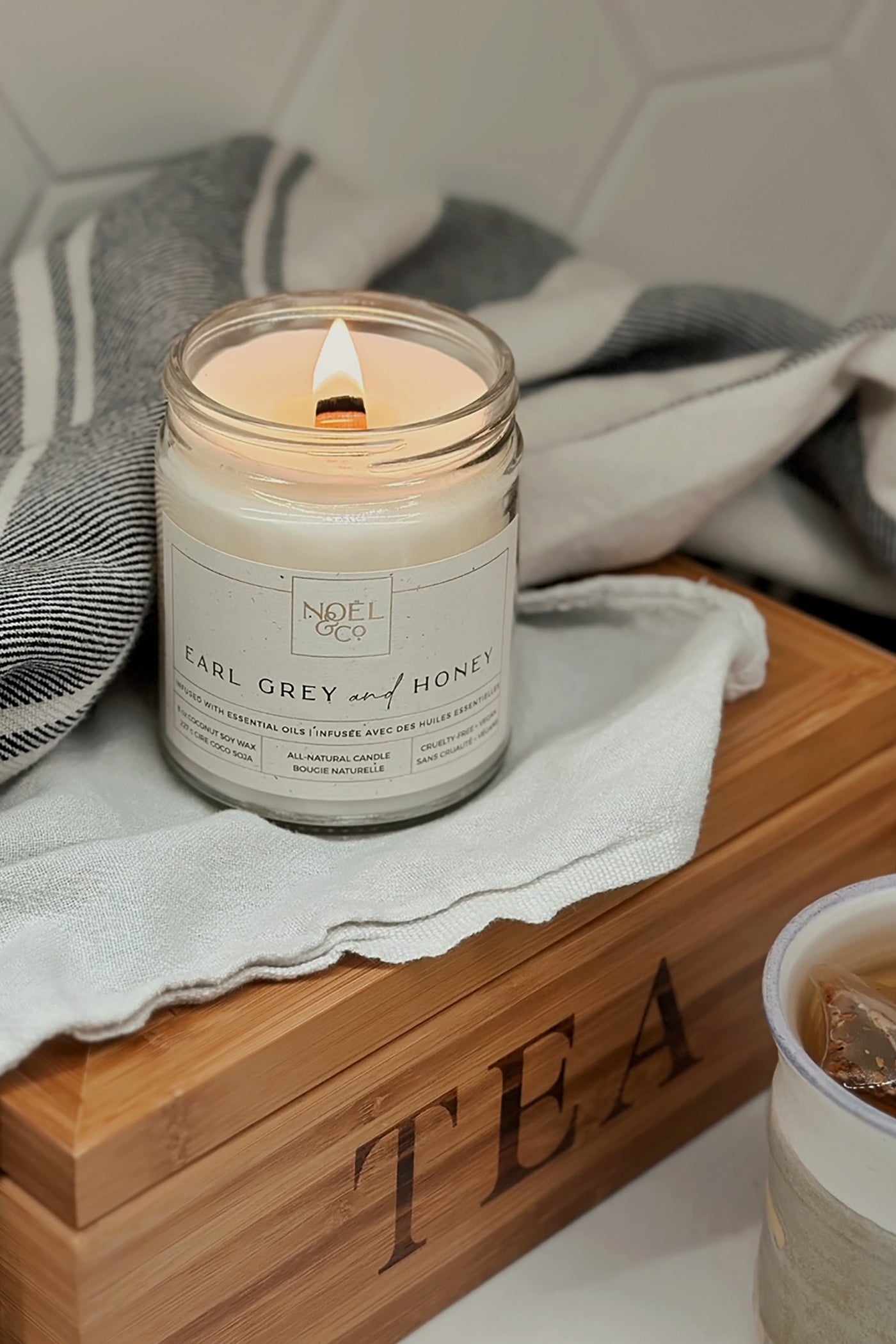 A lit candle in an clear glass jar with a wooden wick sat on top of a box that is labeled “TEA”.