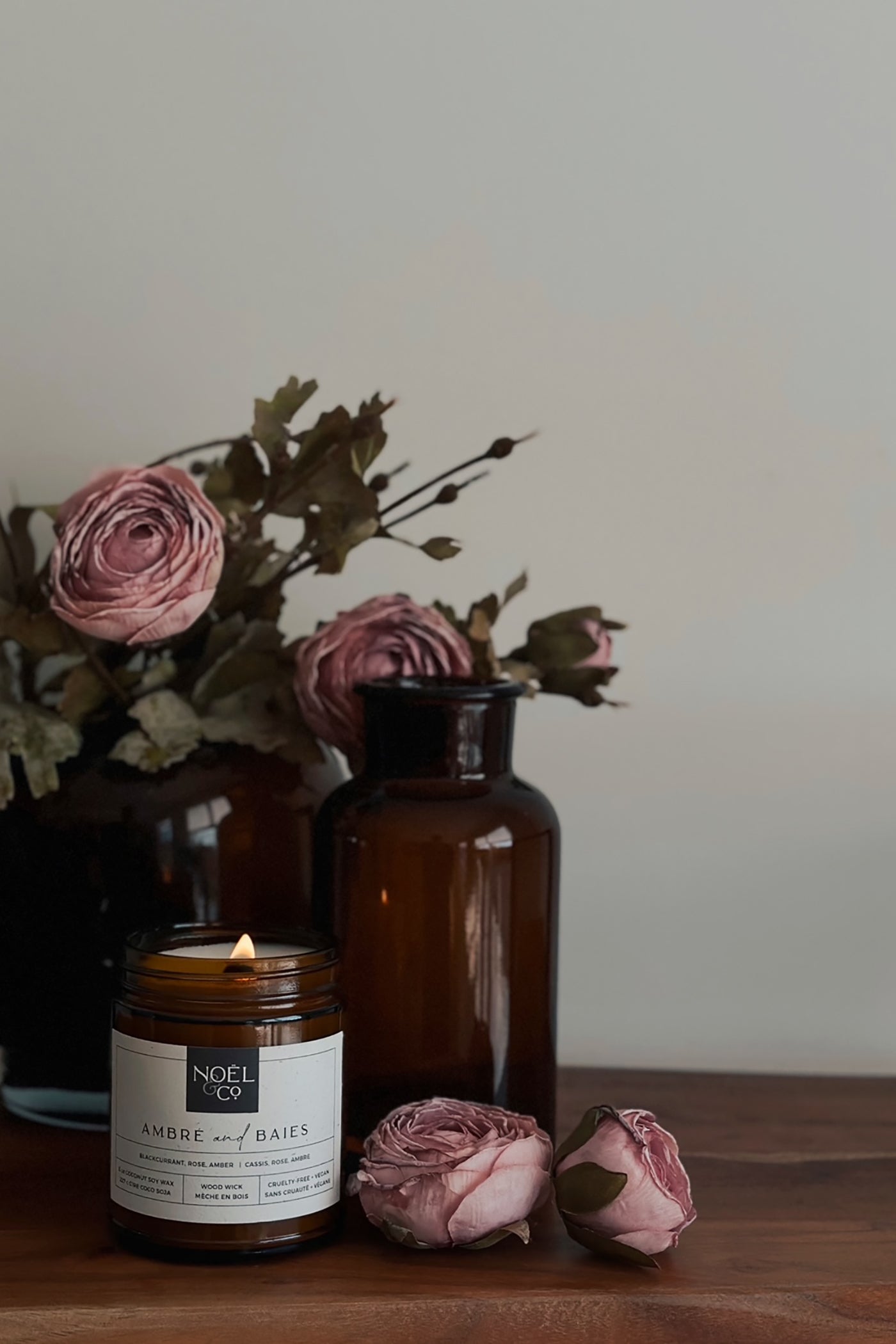 A Noel and Co candle, surrounded by a bouquet of pink roses and brown leaves on a wooden surface.