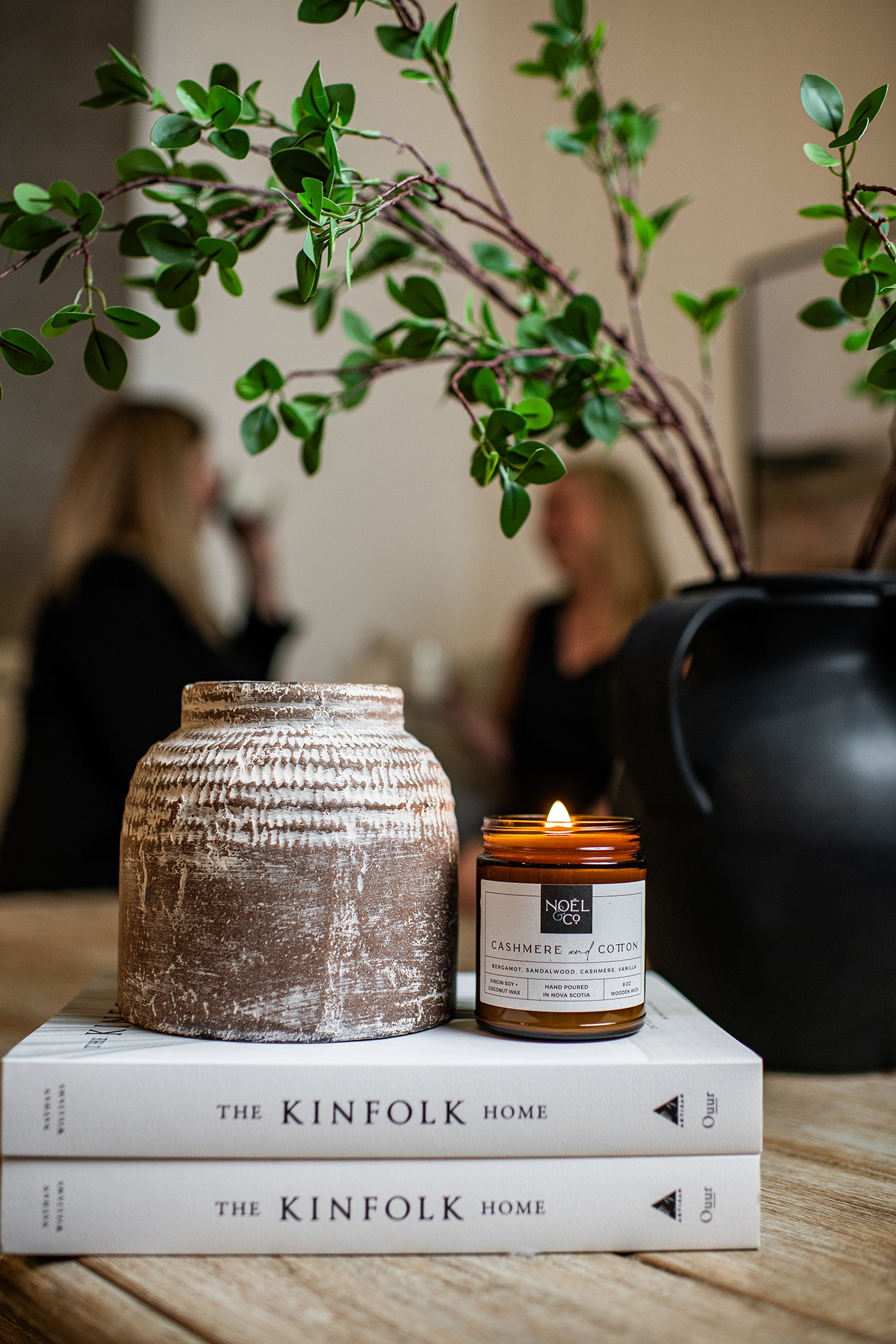 A lit Noel and Co candle in an amber glass jar on two books next to a decorative vase.