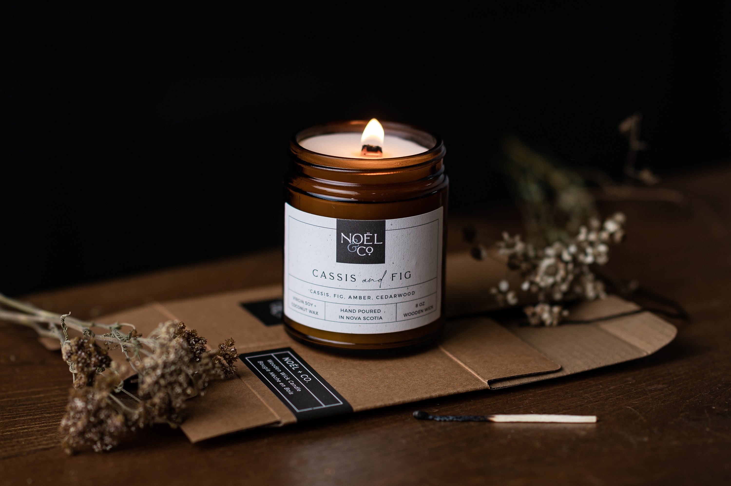 A lit Noel and Co candle in a amber glass jar next to a used match.
