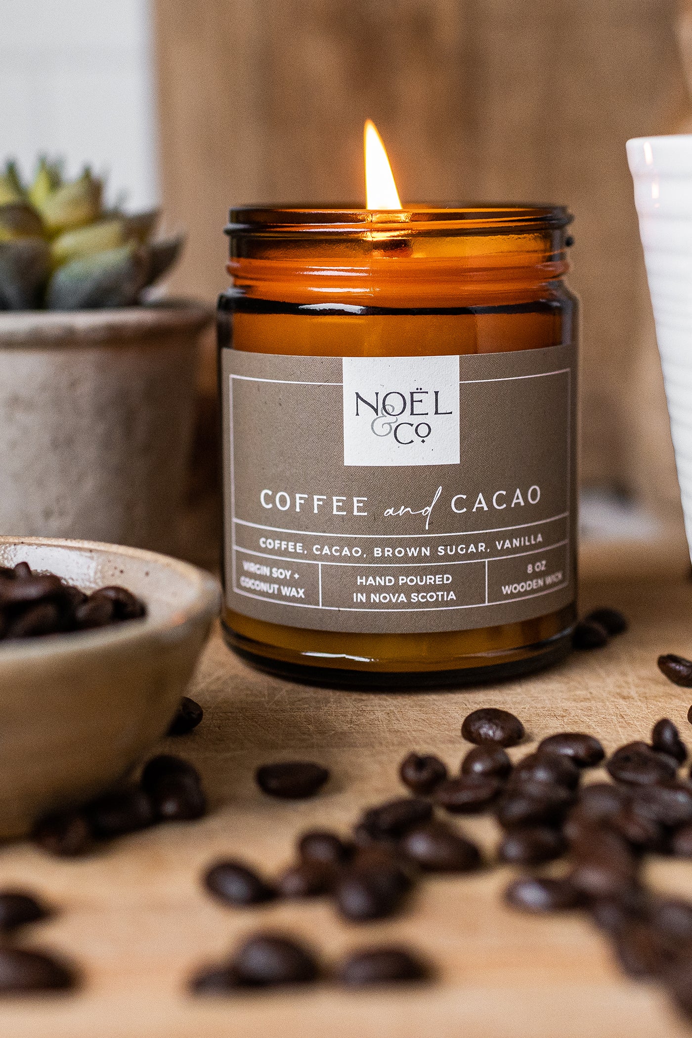 A lit Noel and Co candle in an amber glass jar with a wooden wick and coffee beans on the table around it.