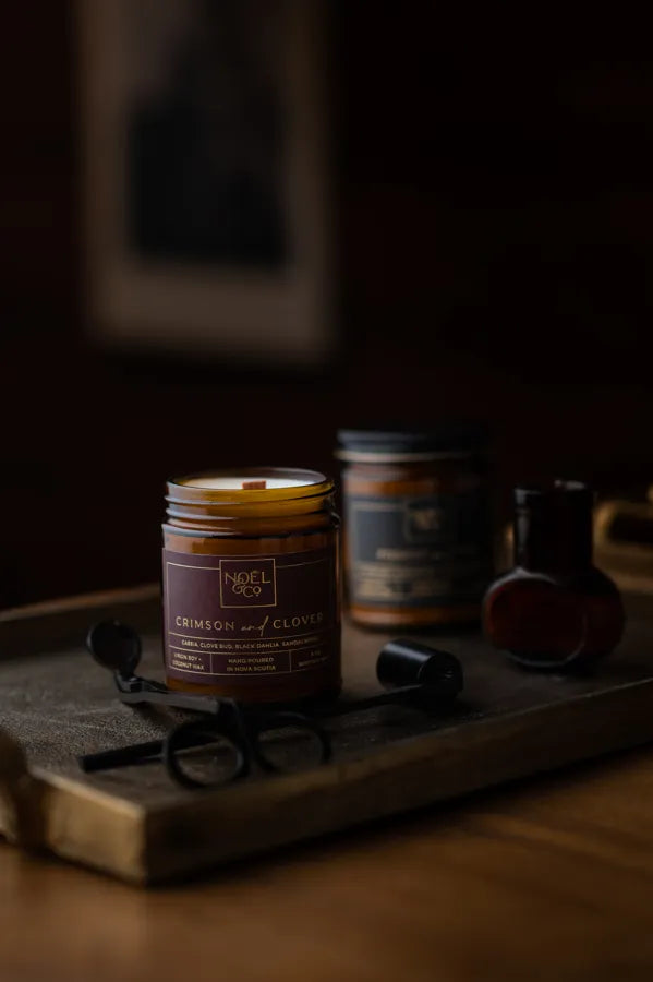 Two Noel and Co candles in amber glass jars. One with a black lid and one without a lid.