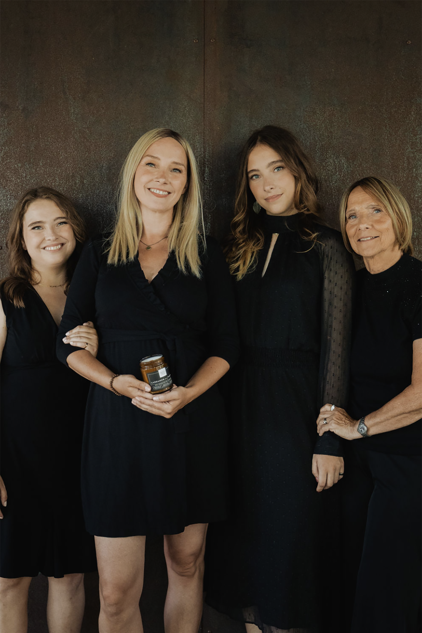 Four women in black attire standing together, with one holding a Noel and Co candle against a dark background.
