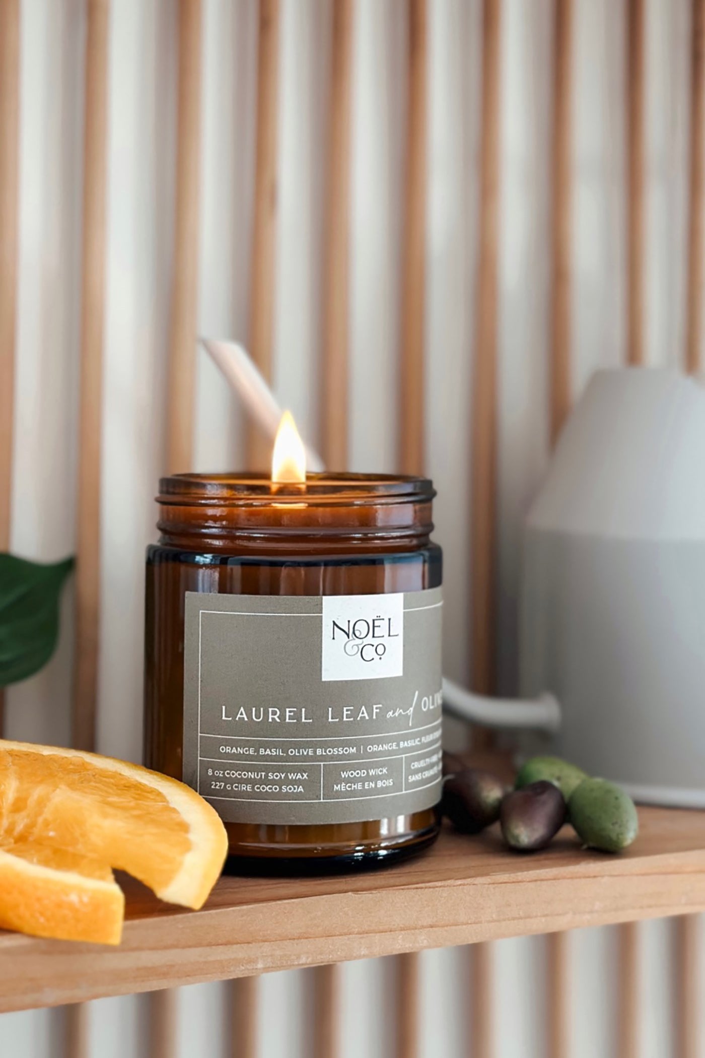 A lit candle with laurel leaf and olive branches in an amber glass jar, labeled 'Noël & Co. Laurel Leaf & Olive', placed on a wooden shelf with a lemon slice and green olive in the foreground.