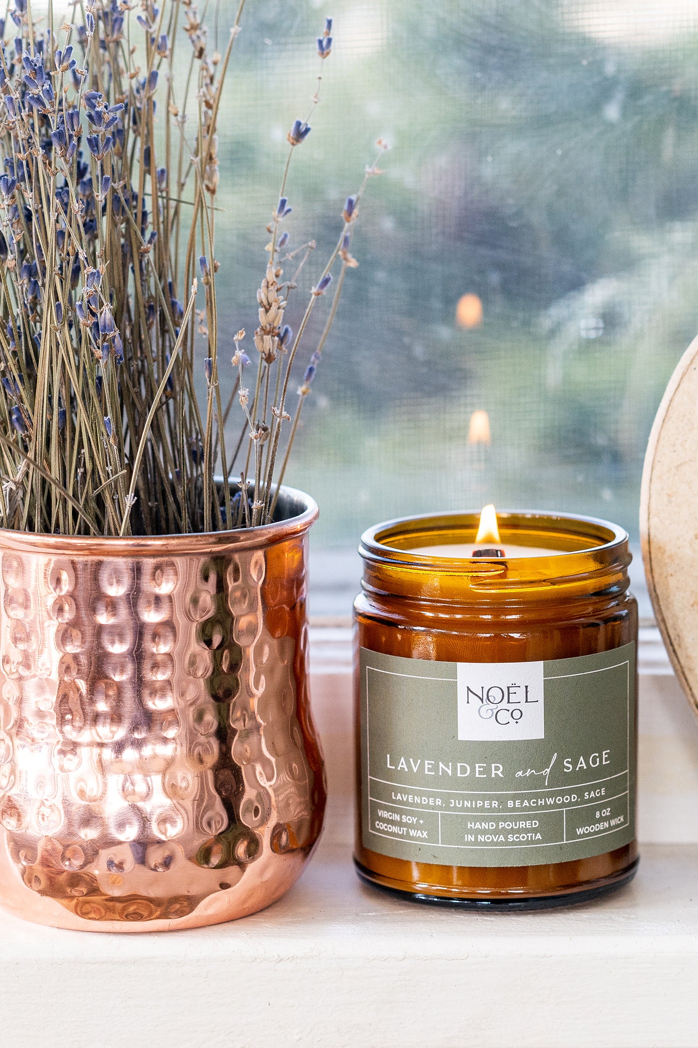 A scented candle labeled 'Lavender & Sage' from Noël & Co, placed next to a copper vase with dried lavender.