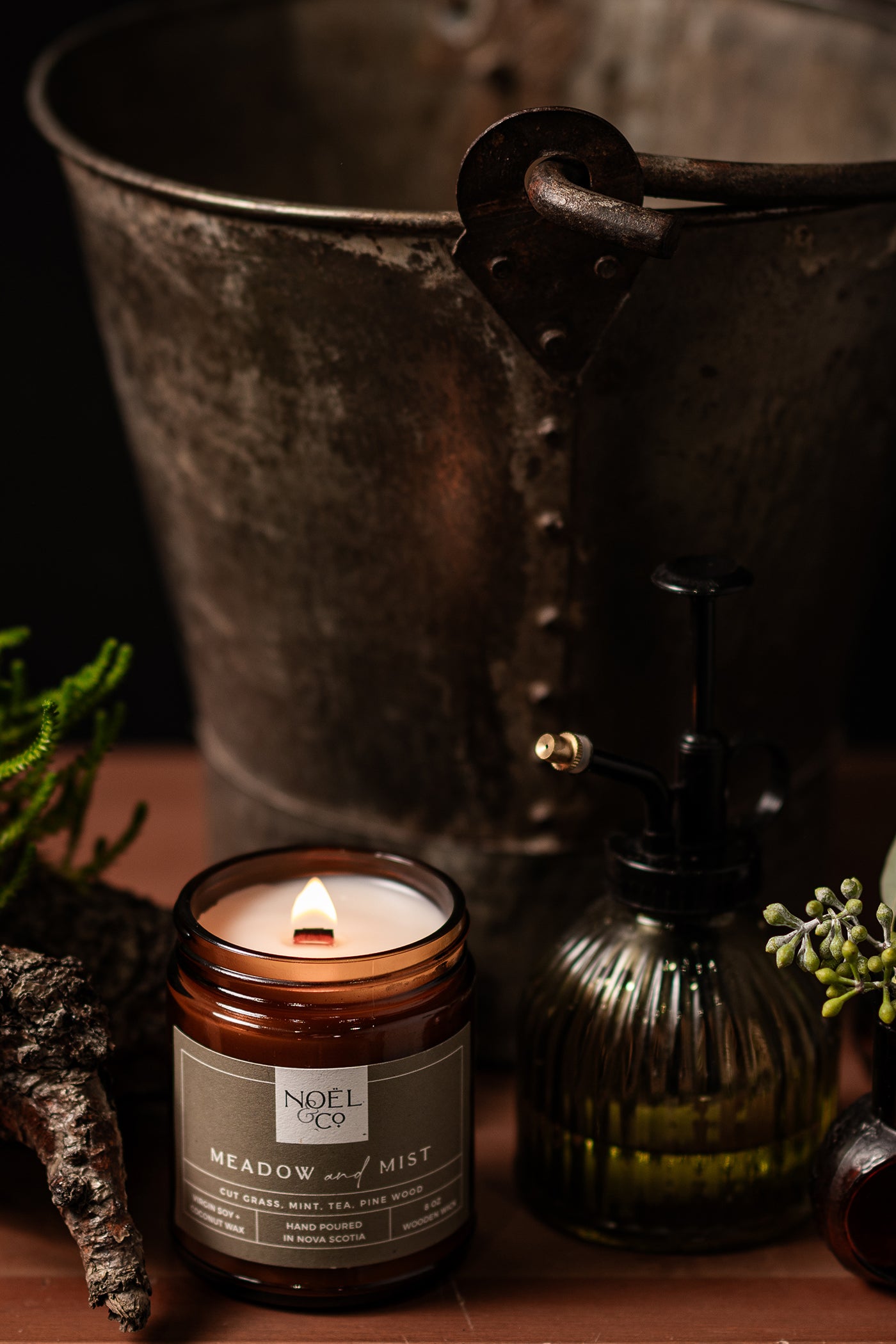 A Noel and Co candle with a label reading 'MEADOW MIST' is lit, emitting a subtle fragrance. It is placed in a rustic metal bucket surrounded by greenery and a glass bottle, emphasizing a connection to nature.