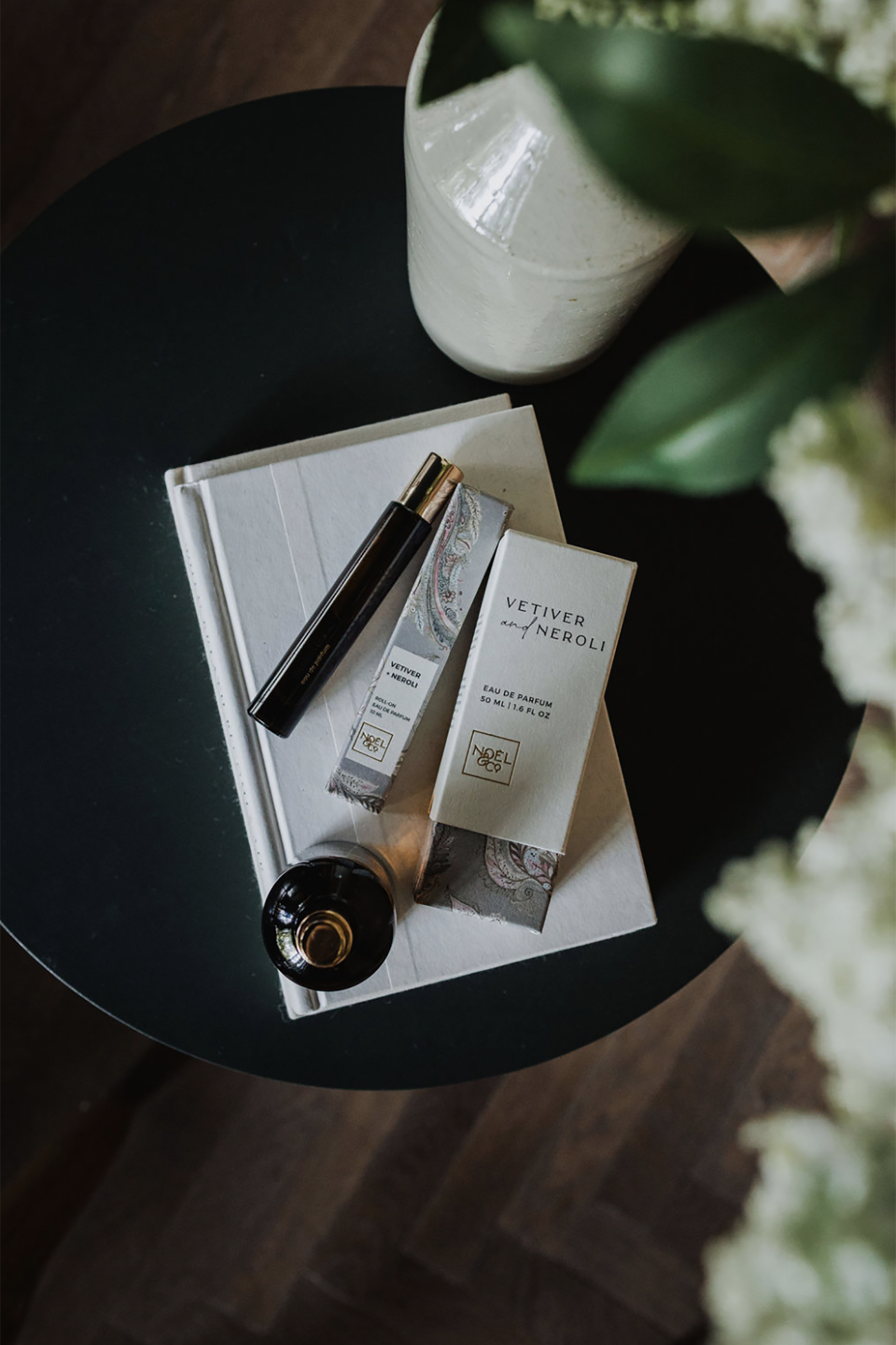 Black round table with Noel and Co Vetiver and Neroli perfume on top, surrounded by green leaves.