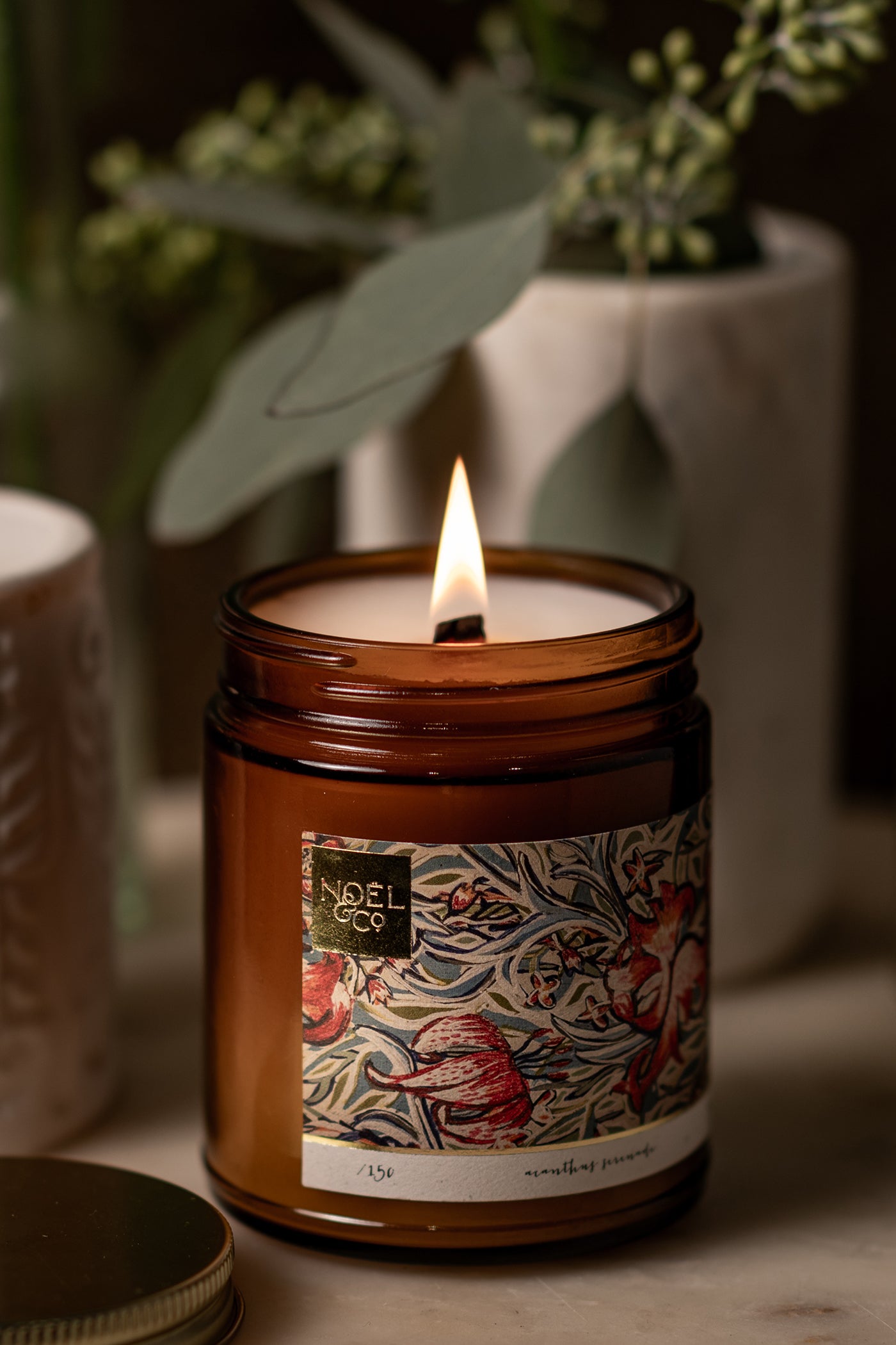 A lit candle in an amber glass jar with a wooden wick and a original art piece featured on the label.