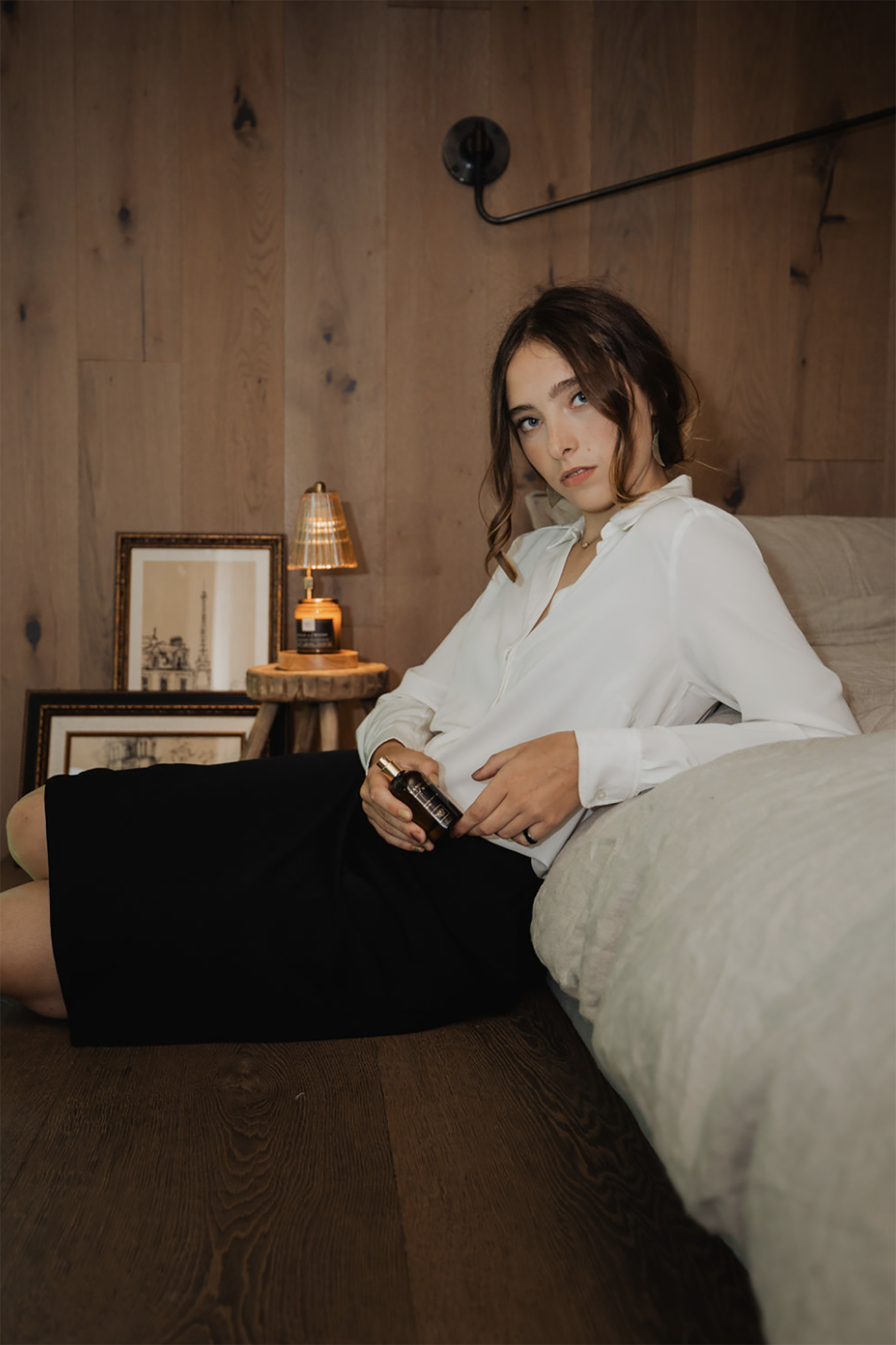 Woman sitting on a bed in a room with a Noel and Co perfume, wooden walls and a lamp