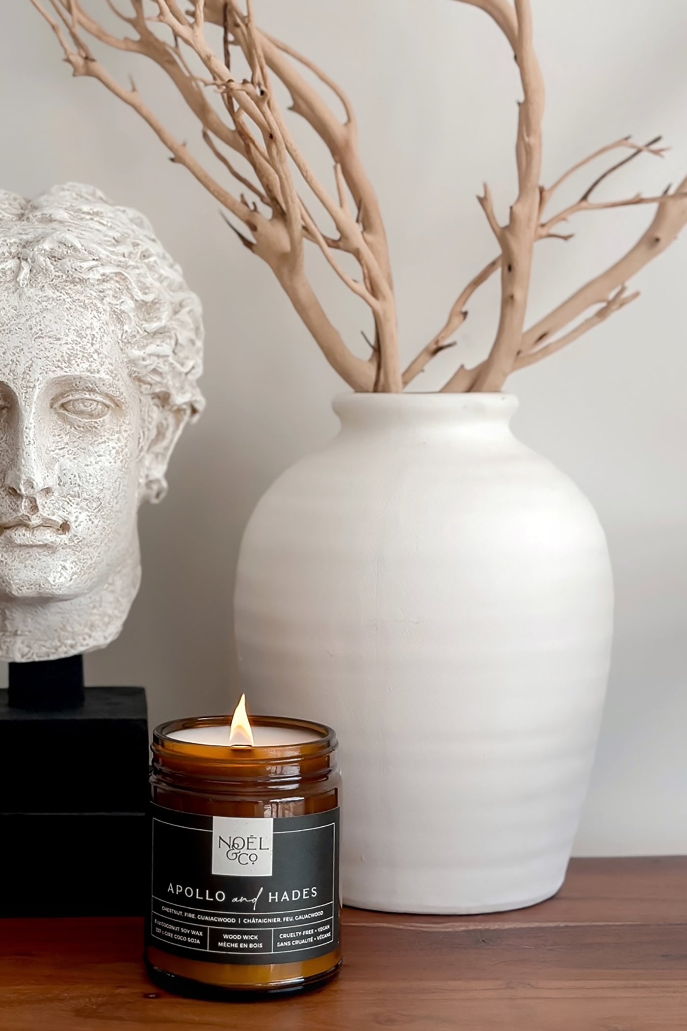 A scented Noel and Co candle labeled 'Apollo & Hades' is lit, placed on a wooden surface with a white vase containing dried branches in the background, and a classical bust sculpture to the left.