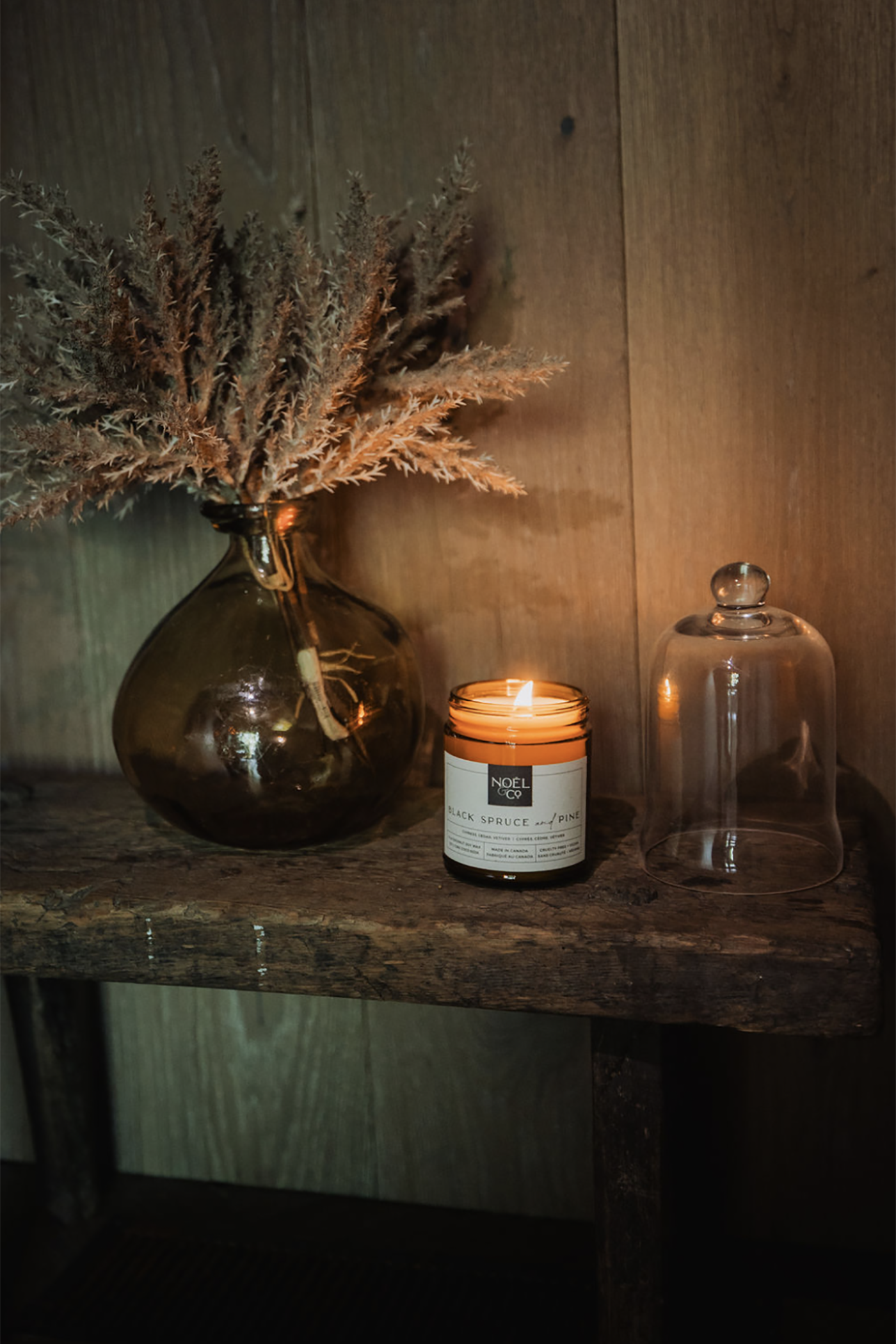 A Noel and Co candle with a label on a wooden surface next to a vase with dried plants and a glass jar.