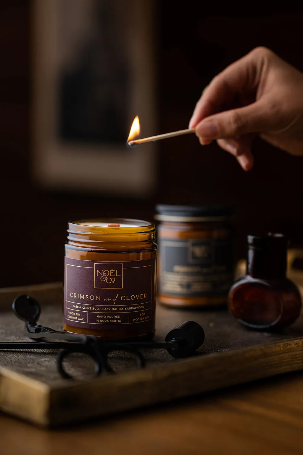 Two Noel and Co candles in amber glass jars. One with a black lid and one without a lid. A hand holding a lit match above the candle without the lid.