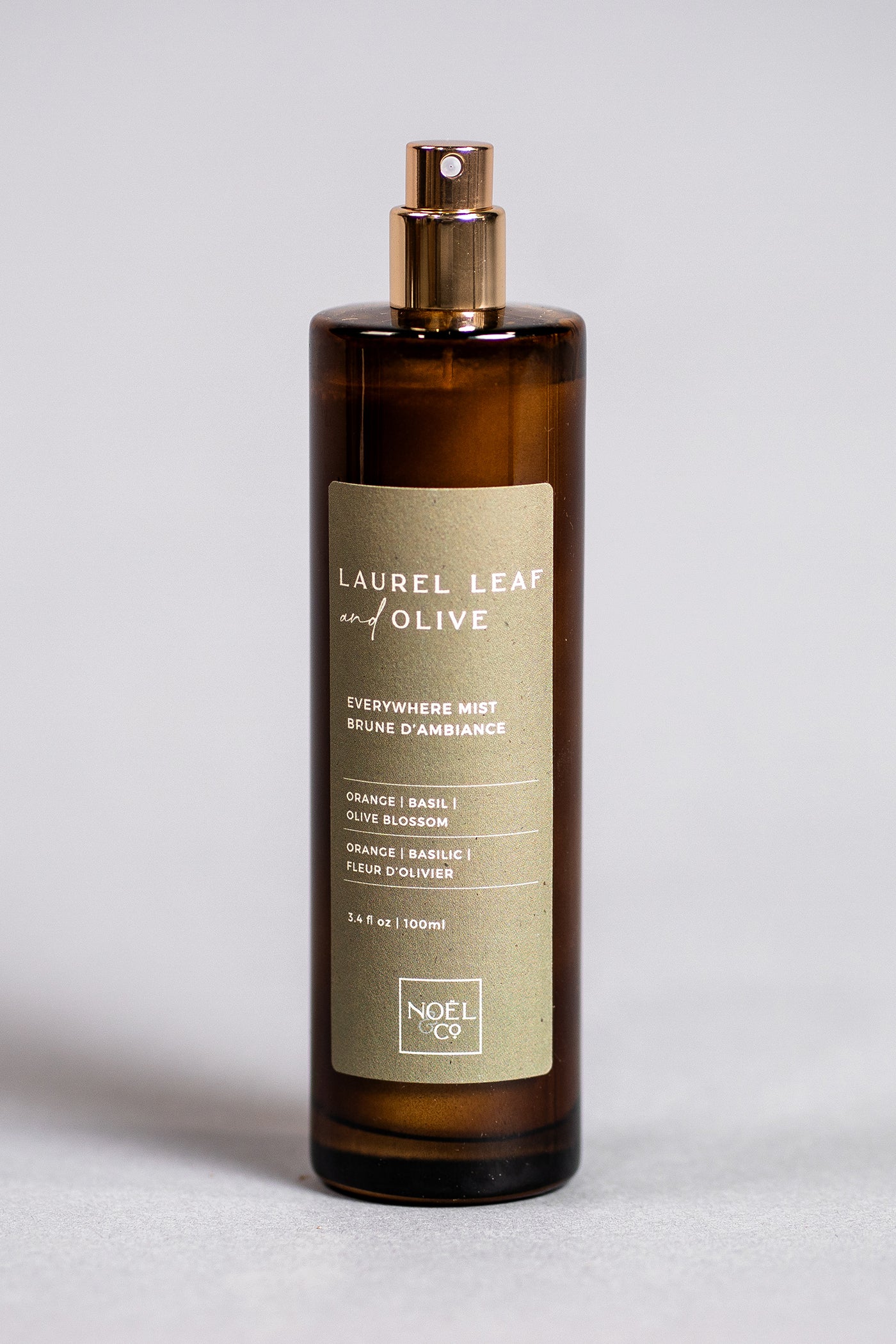 An amber glass Noel and Co everywhere mist with a gold lid, labeled 'LAUREL LEAF OF OLIVE' along with other product details.