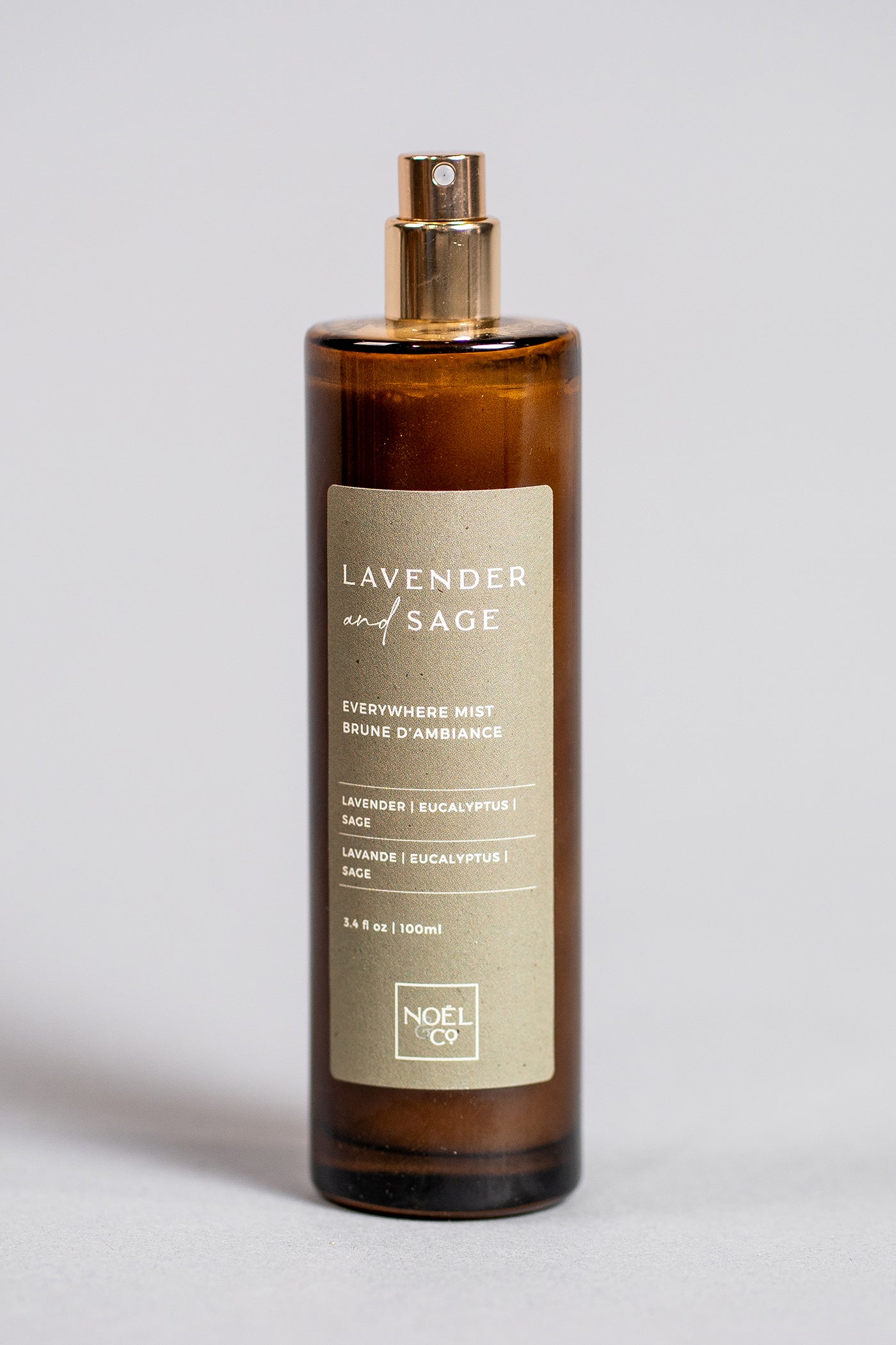 A brown glass everywhere mist bottle with a label that reads 'Lavender & Sage' and 'Noël & Co'. The cap of the jar is gold-colored.