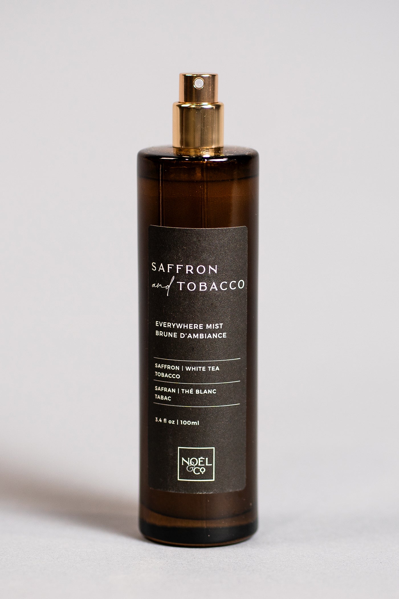 A tall, slender bottle of Noel and Co Saffron & Tobacco everywhere mist with a gold-colored cap, labeled in a simple, elegant font against a white background.