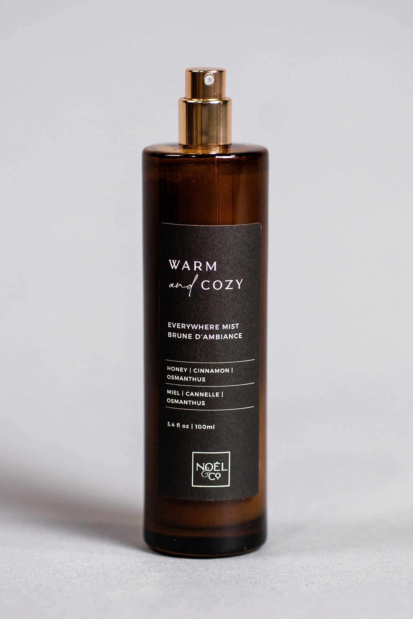 A bottle of Noel and Co 'Warm & Cozy' everywhere mist with a dark amber tint and a gold spray nozzle. The label features a black background with white text detailing the scent story and key notes.