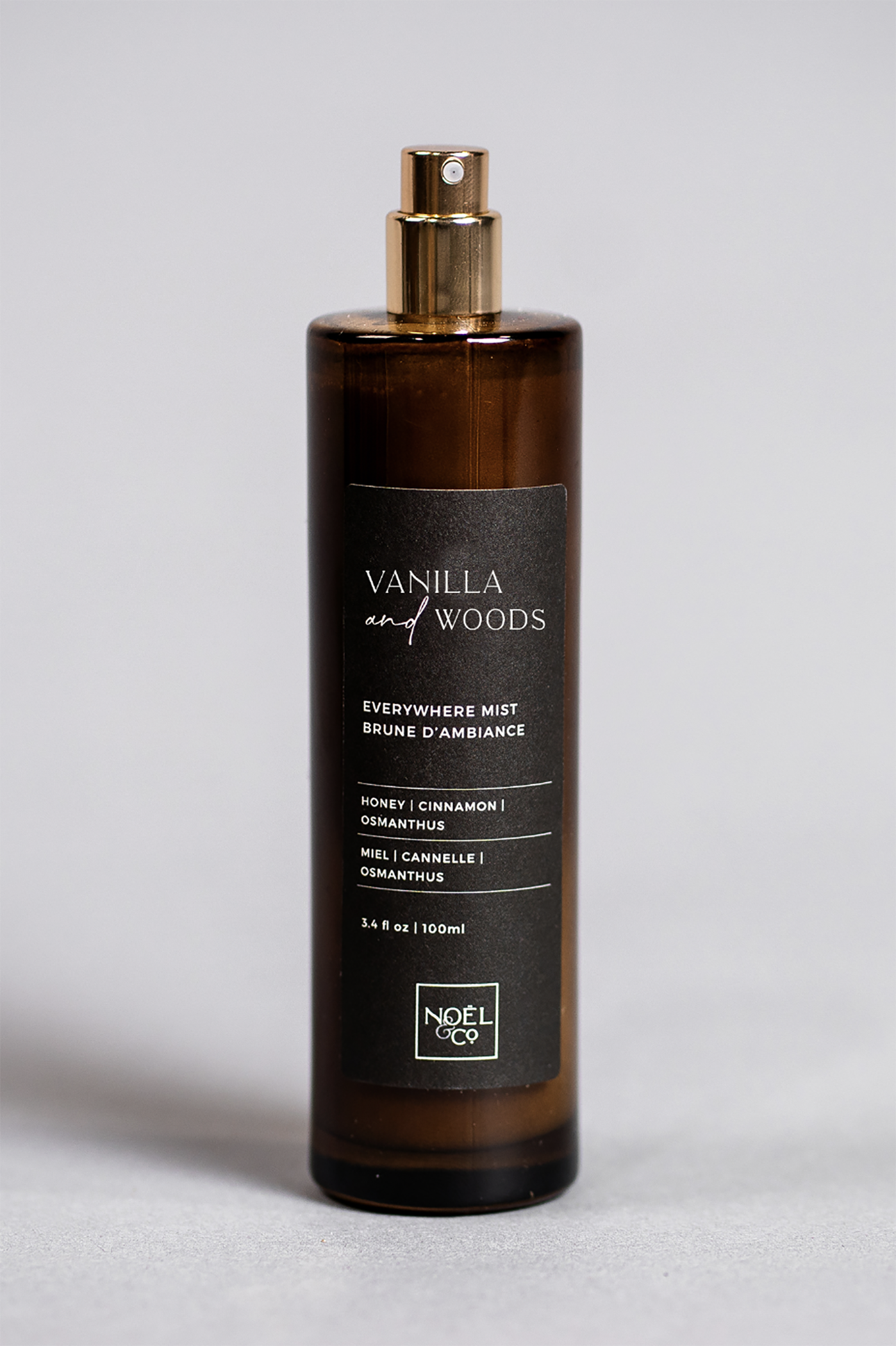 A bottle of 'Vanilla & Woods' room mist with a brown tint and a gold-colored spray nozzle. The label is black with white and gold text.