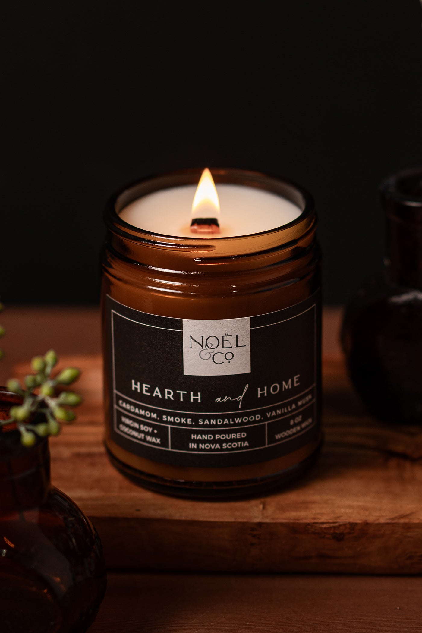 A lit candle with a smoky aroma, placed on a wooden surface, with a label that reads 'HEARTH & HOME' from Noel & Co.