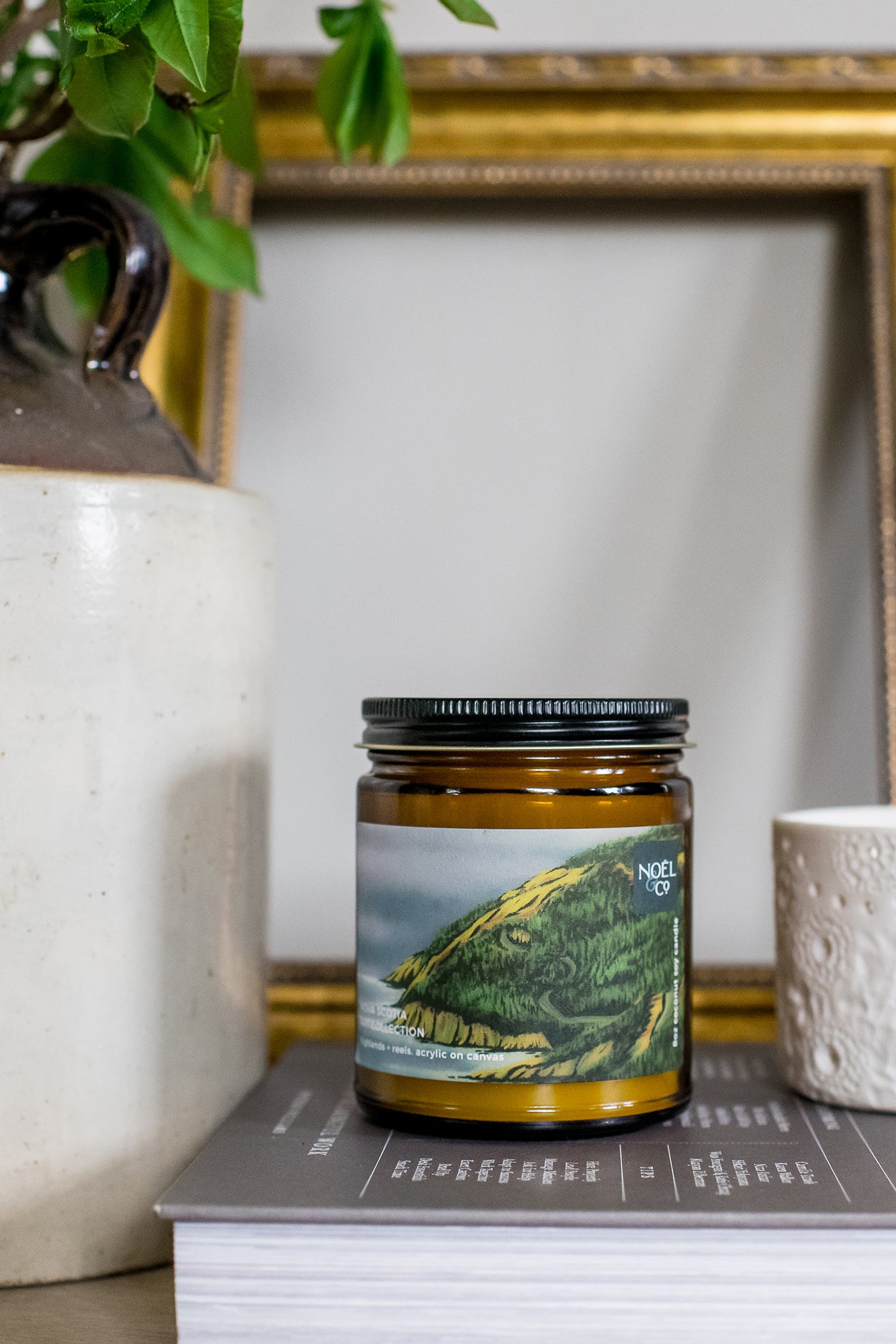 A Noel and Co candle with a label depicting Cape Breton Nova Scotia, placed on a book next to a cup and a plant, with a framed picture in the background.