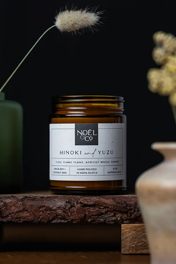 A Hinoki & Yuzu scented candle by Noël & Co. placed on a wooden surface with a dark background, decorative vase, and a sprig of dried plant in the foreground.