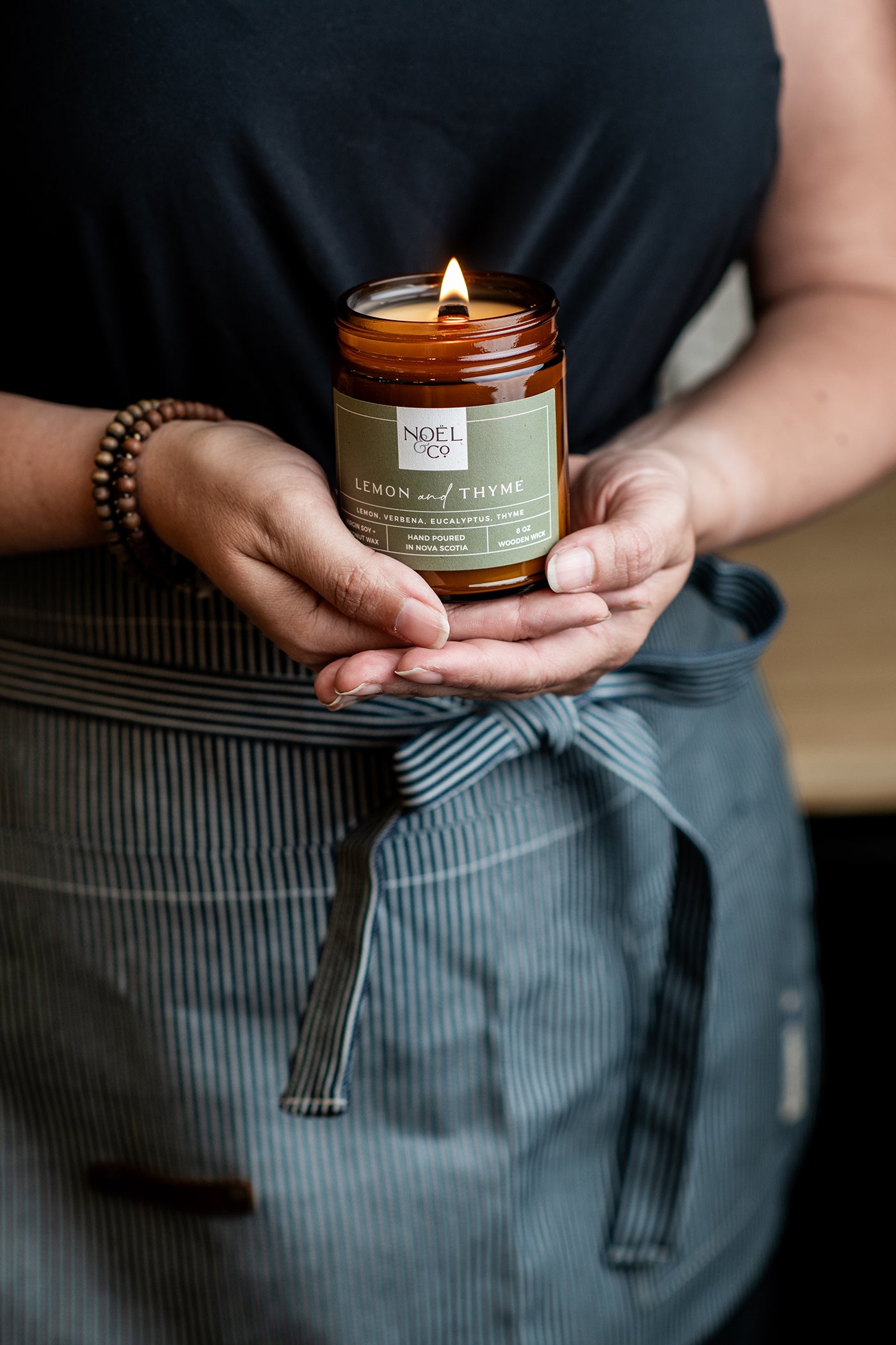 A person holding a lit Noel and Co candle with Lemon & Thyme label, providing a warm glow.