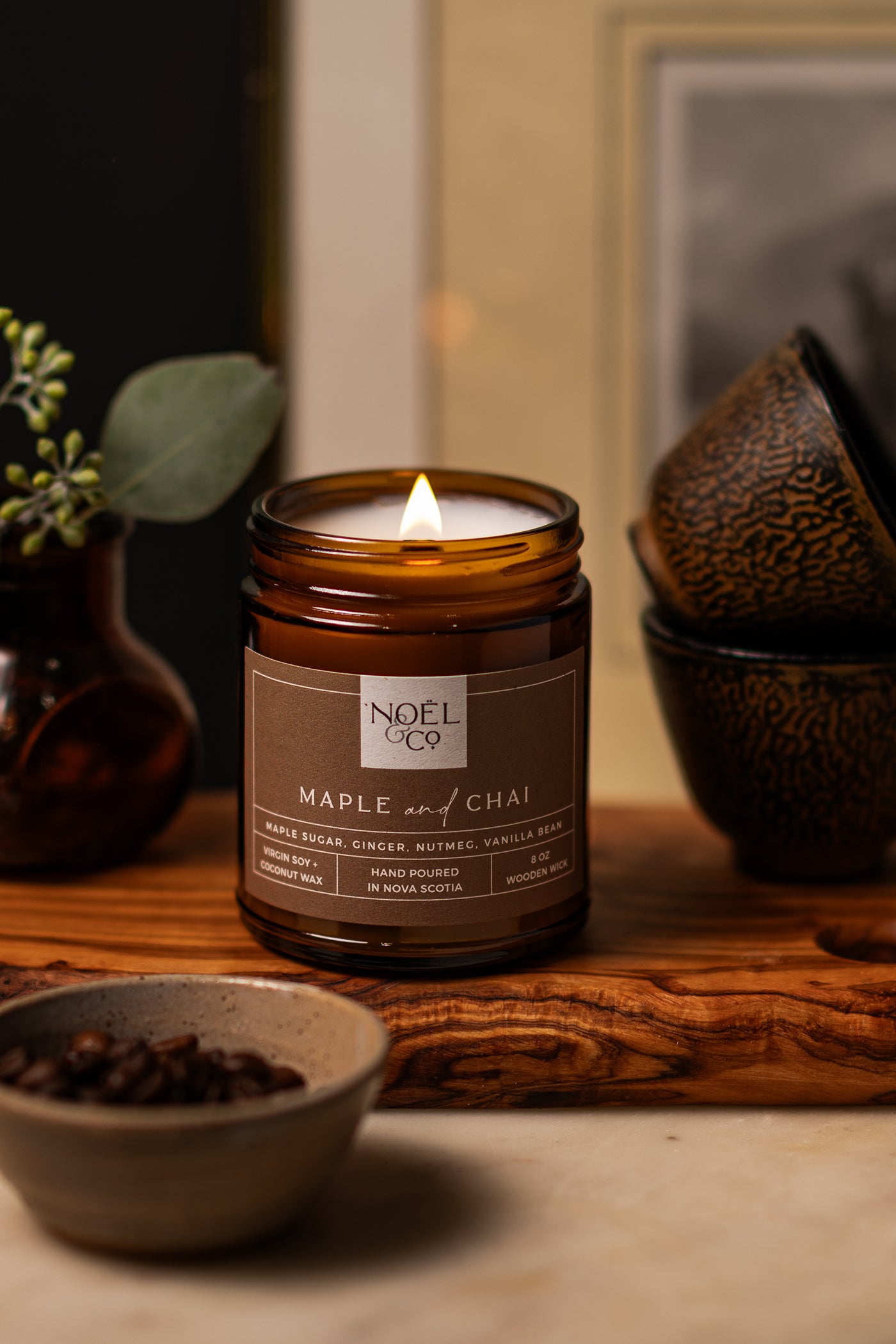 A lit Noel and Co candle with 'Maple & Chai' written on its label, placed on a wooden surface with bowls around it.