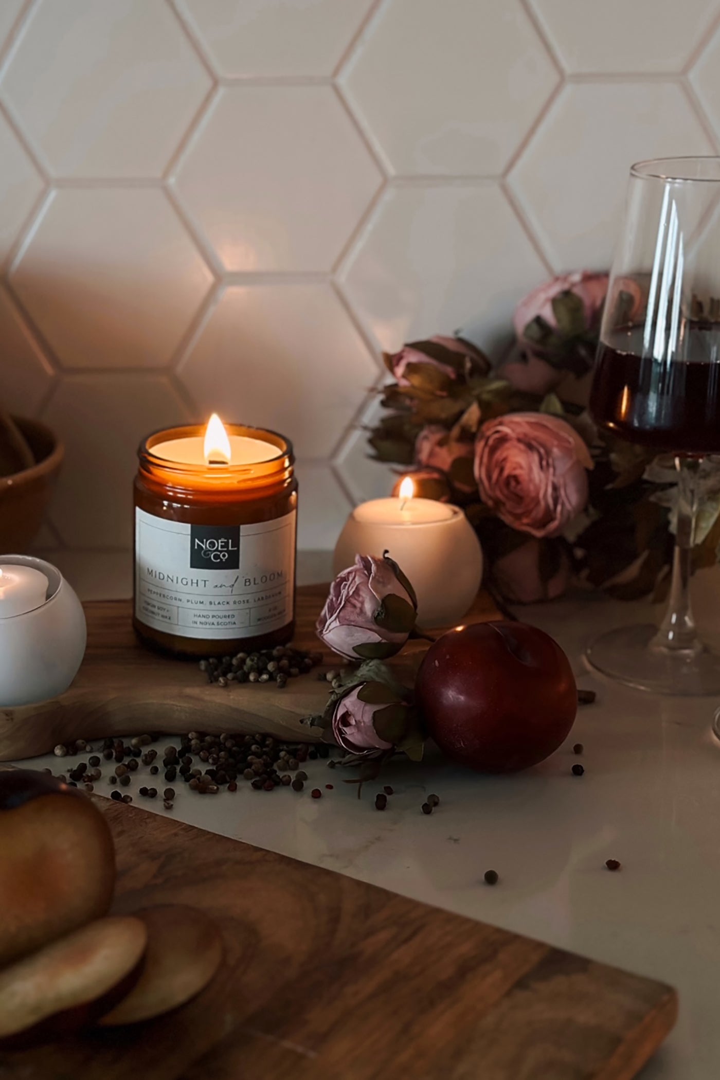 A lit Noel and Co candle in an amber glass jar with a white label, surrounded by decorative items on a table, with a muted background featuring a floral arrangement and a glass of wine.