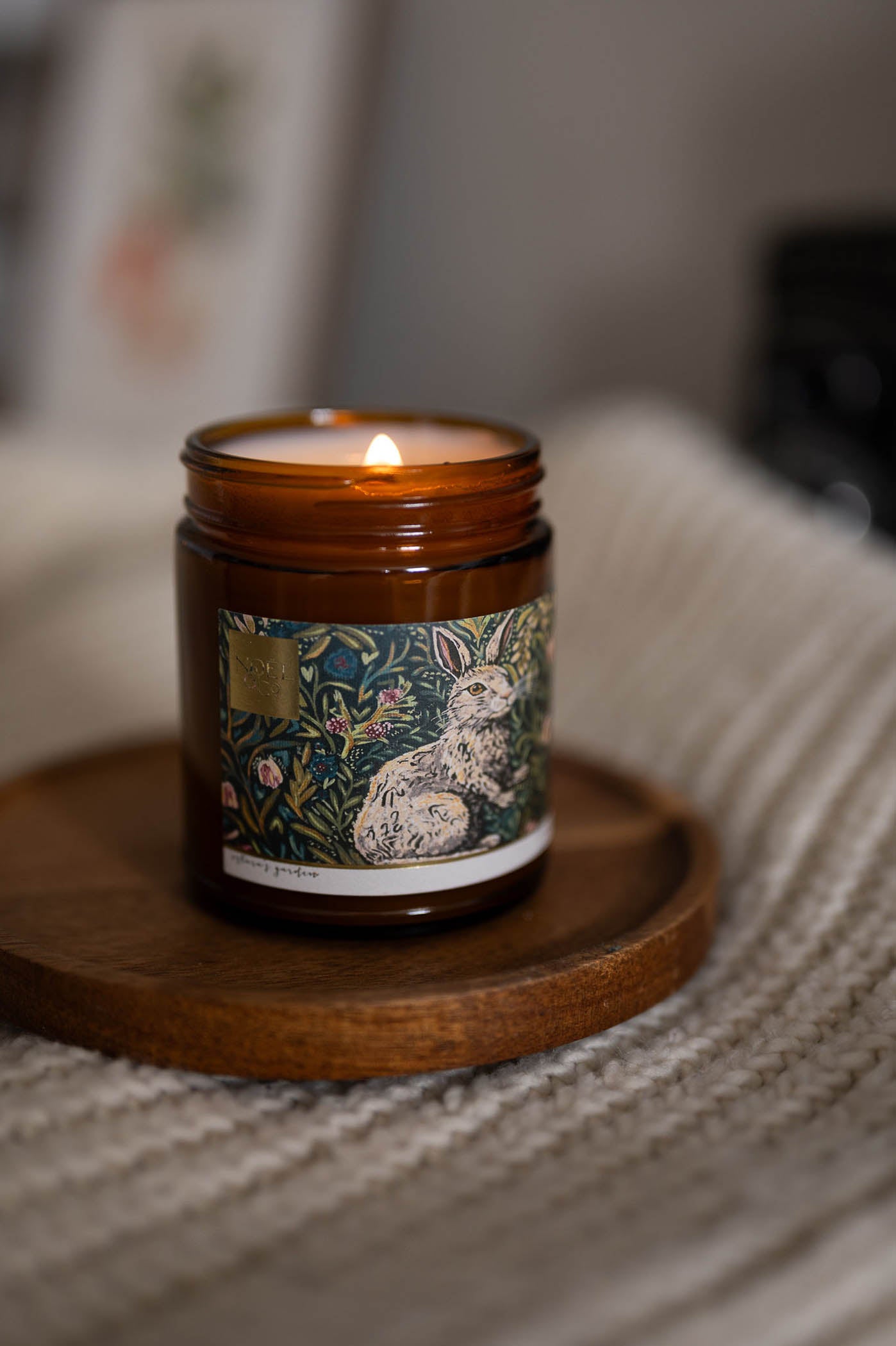 A lit Noel and Co scented candle with a floral and rabbit design on the label, placed on a wooden surface.