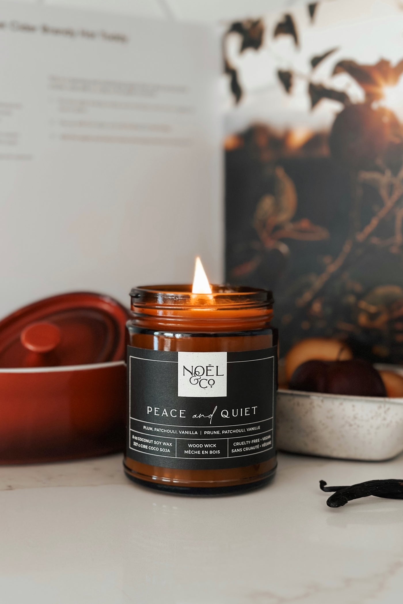 A lit candle with the label 'NOÉL & CO. PEACE & QUIET' in an amber glass jar, surrounded by a cozy setting with books and a warm light.