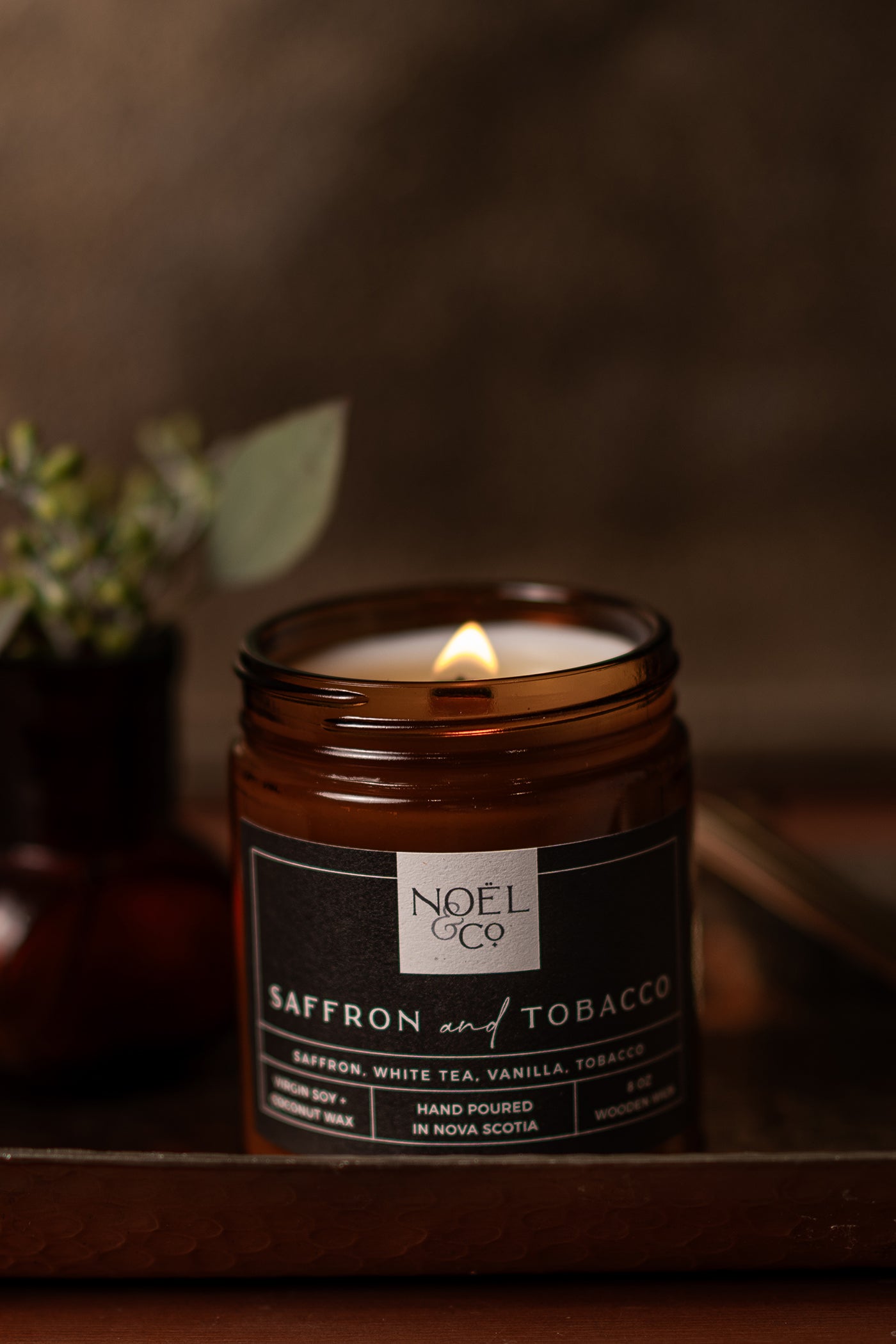 A lit Noel and Co candle in an amber jar with a label reading 'Saffron & Tobacco', with a dark background.