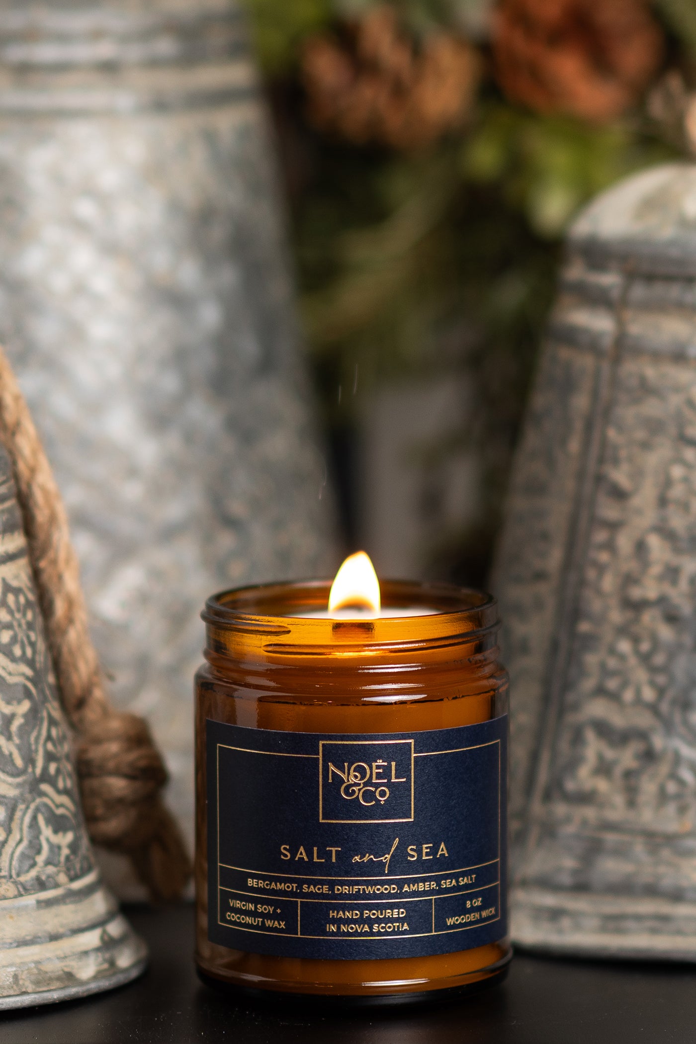 A lit Noel and Co candle with a label that reads 'SALT & SEA', placed on a surface with decorative items in the background.