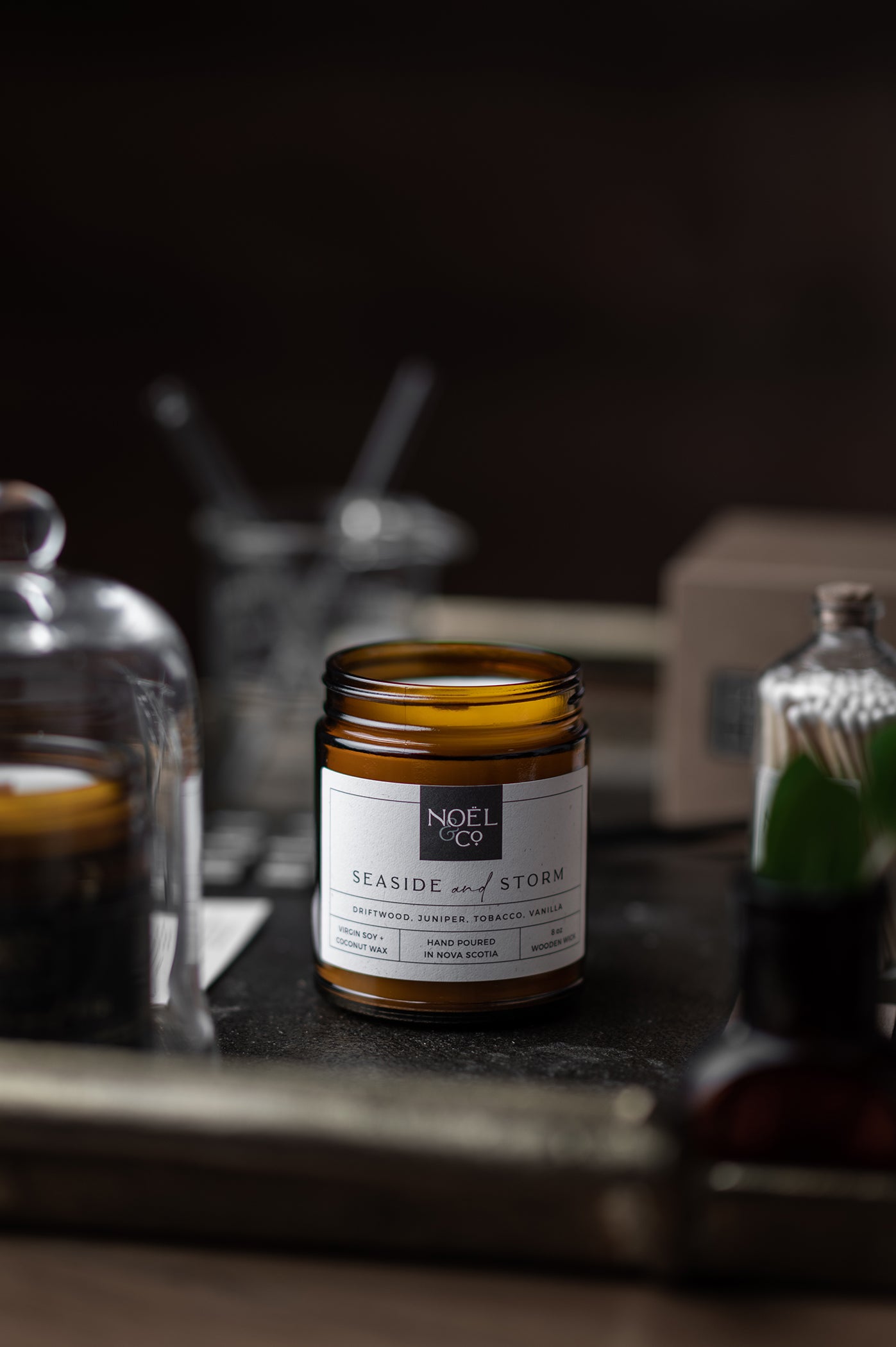 An amber glass jar Noel and Co candle with a white label on it, placed on a metal surface with a dark background. There are blurred objects in the background.