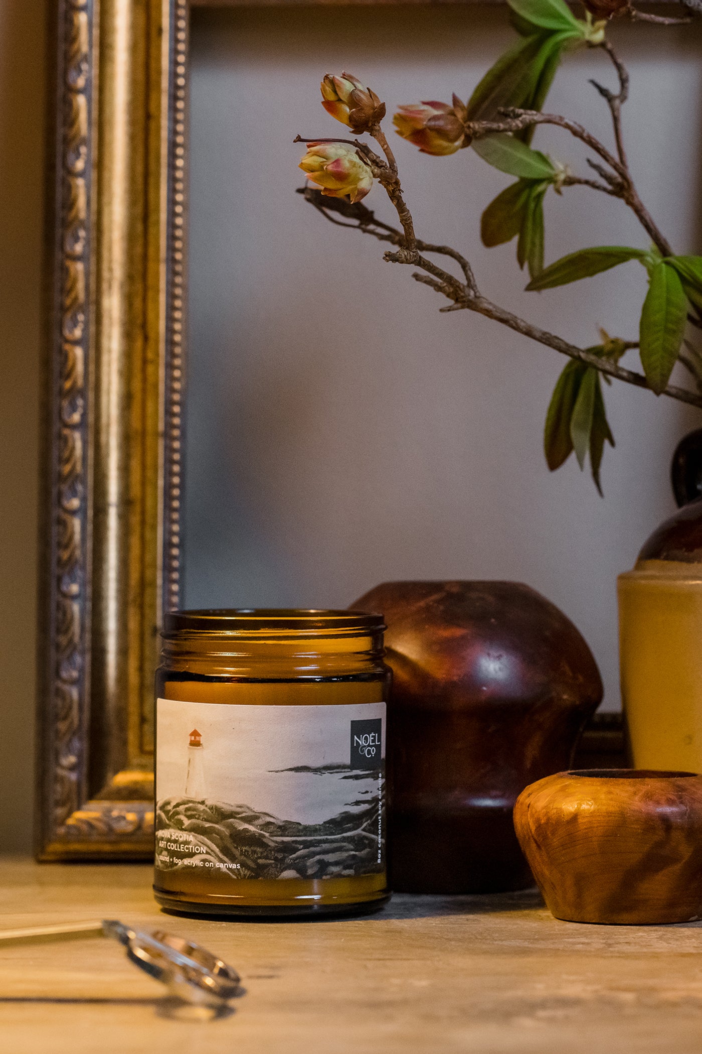A Noel and Co candle with a label of a painting of Peggy's Cove, placed on a wooden surface, with a framed picture and a plant in the background.