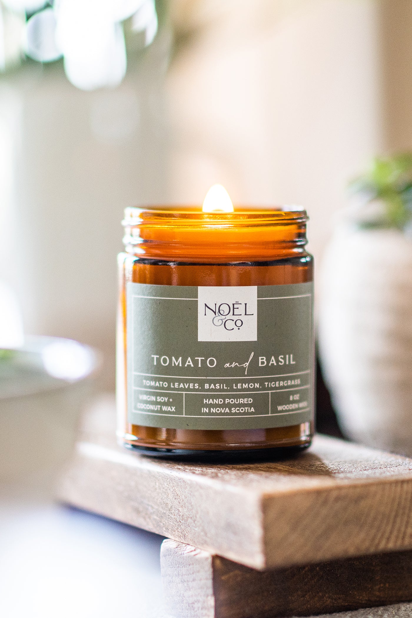 A scented candle labeled 'NOEL & CO TOMATO & BASIL' in an amber glass jar, placed on a wooden surface.