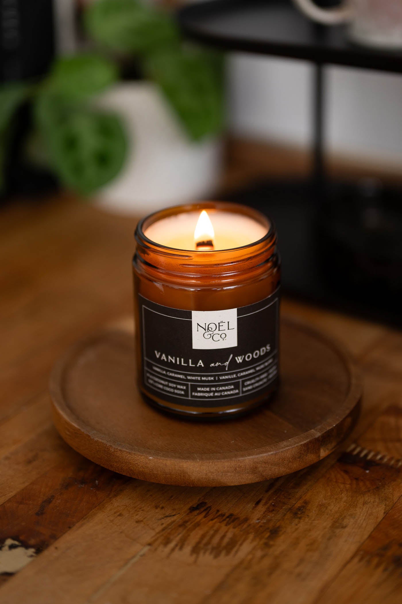 A lit Noel and Co candle with the scent of vanilla and woods, placed on a wooden surface.