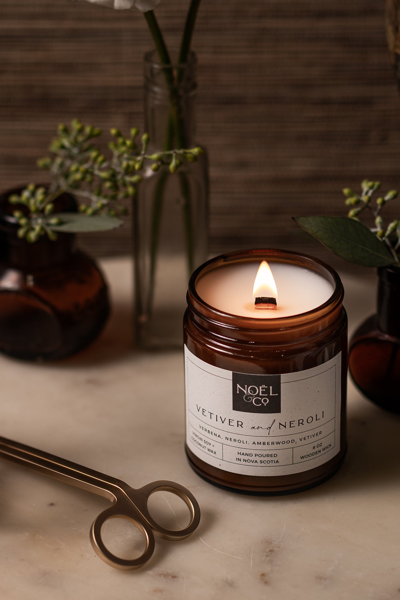 A lit Vetiver & Neroli scented Noel and Co candle in an amber glass jar with a white label, placed on a marble surface with a pair of wick trimmers and decorative plants in the background.