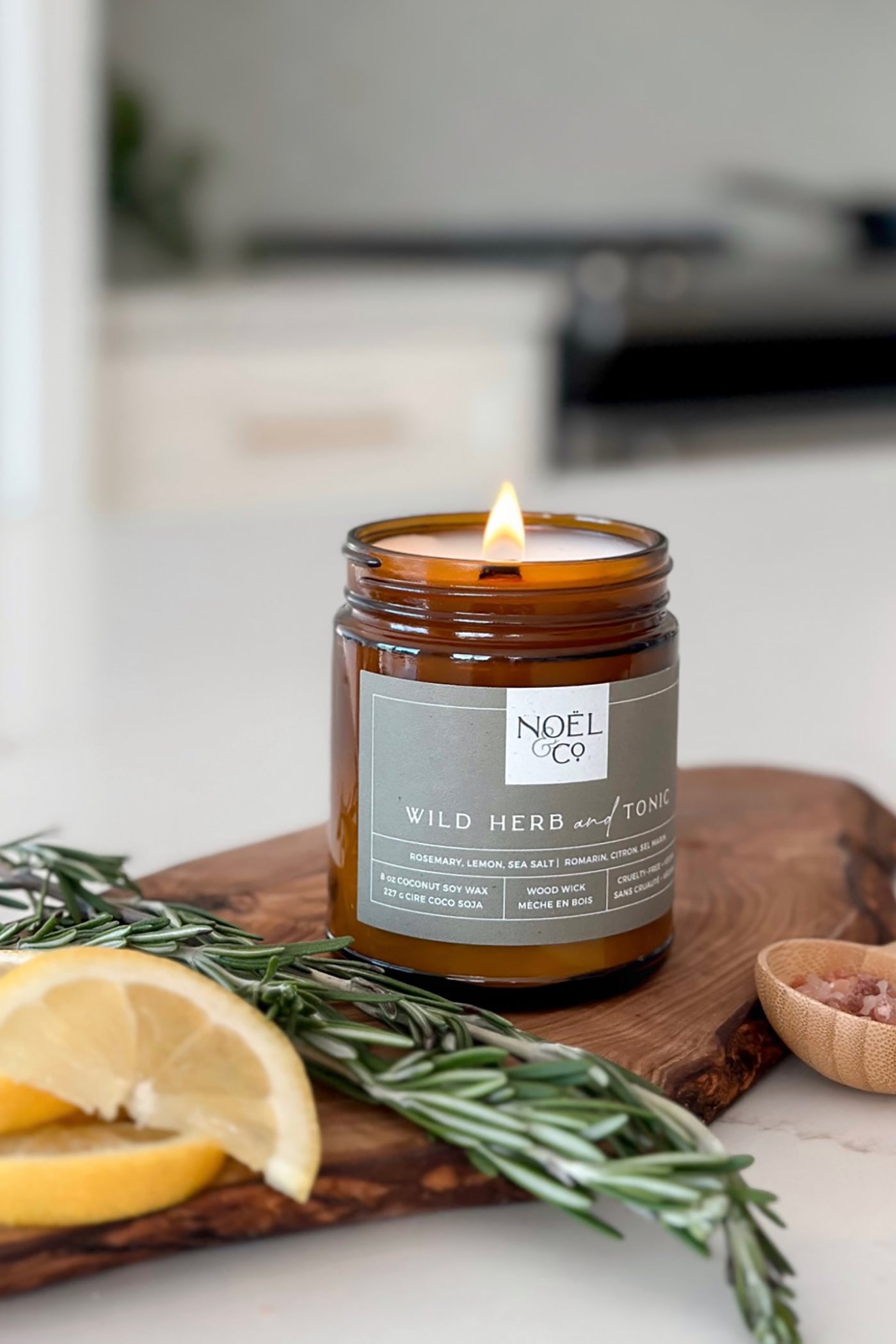 A candle with a label 'Noël & Co. Wild Herb & Tonic' placed on a wooden board, surrounded by a lemon slice, rosemary sprigs, and sea salt.